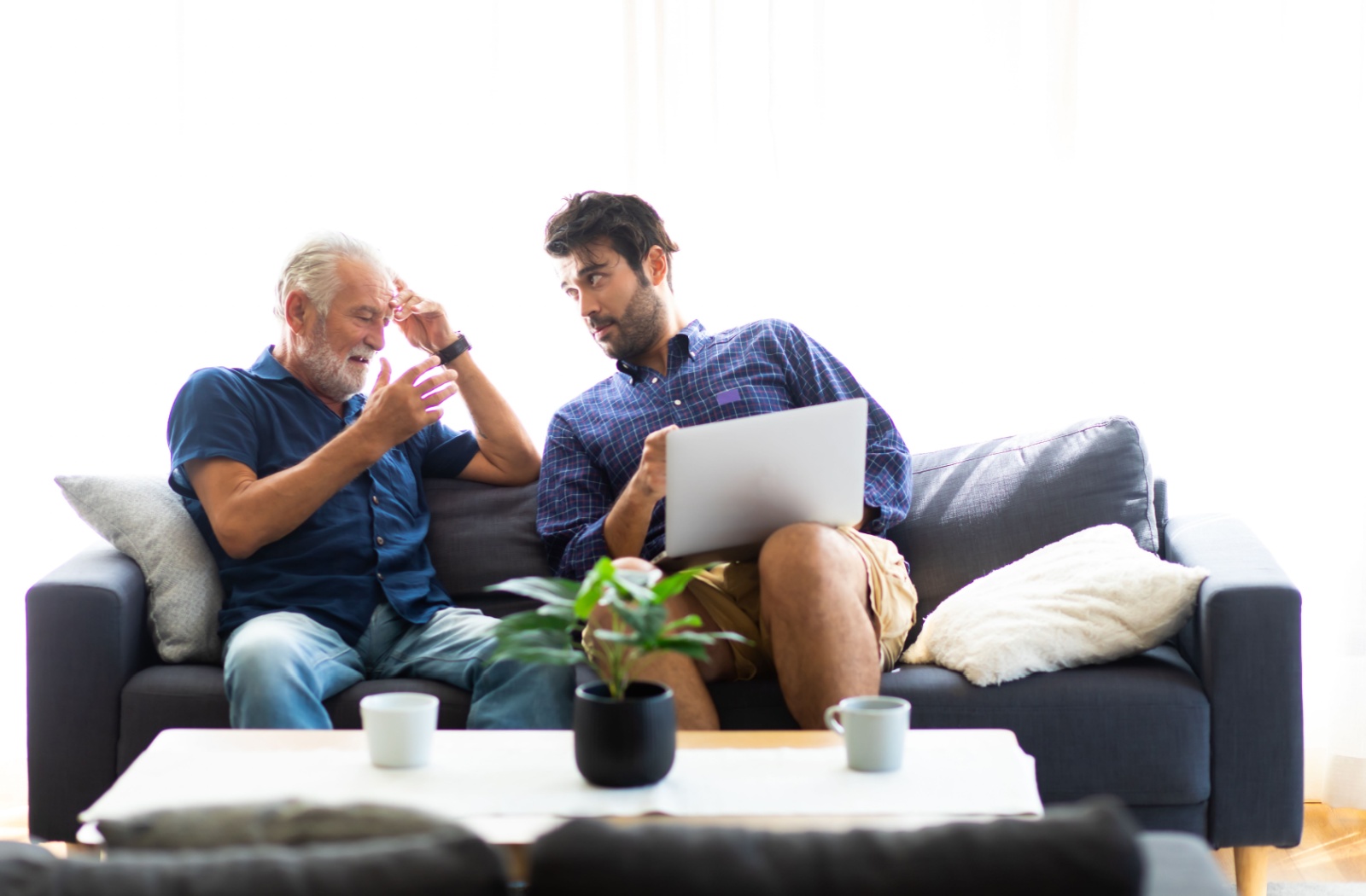 An adult son and elderly father are in a discussion, looking at a laptop over coffee.