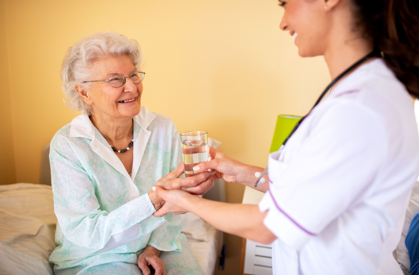 A caregiver in skilled nursing handing a smiling senior woman a glass of water.
