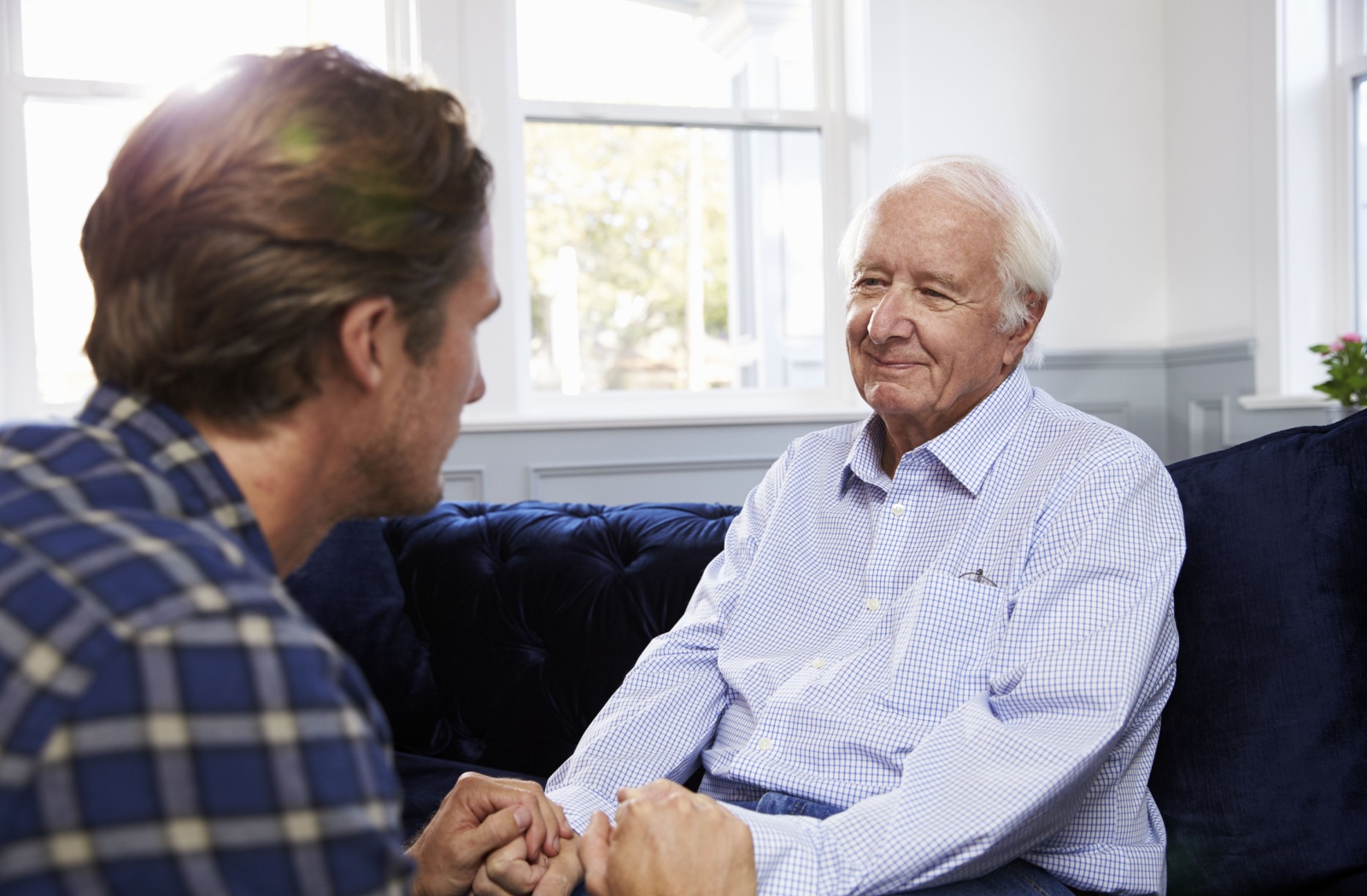 An adult son holding hands with his smiling senior father making eye contact during a conversation.