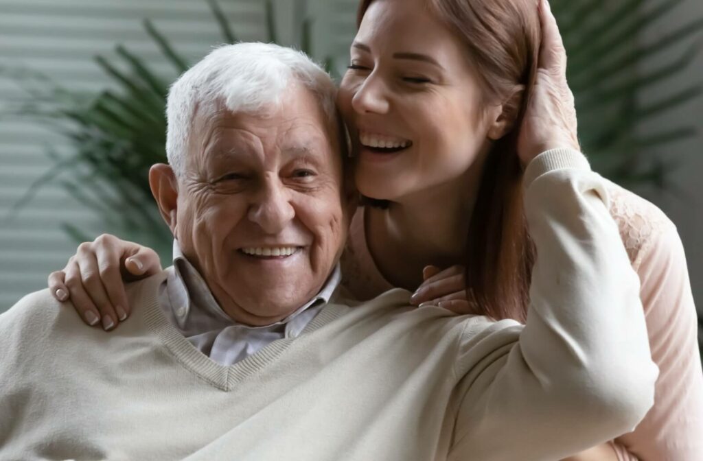 A senior man laughing while his smiling adult daughter hugs him from behind.