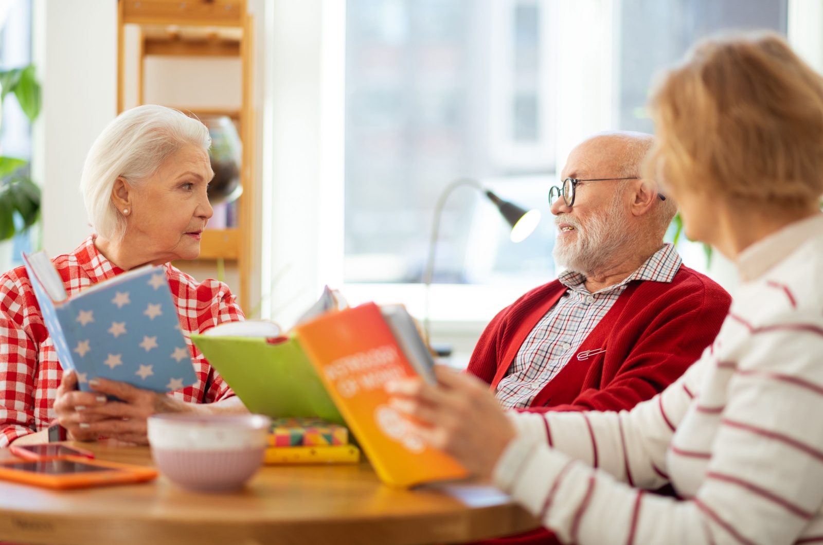 A group of seniors in a book club sitting at a table with books in their hands having a discussion.