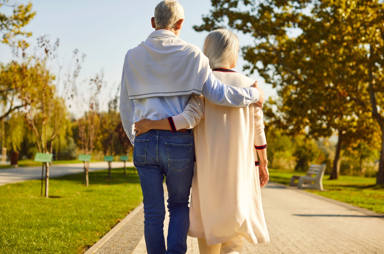 A view of a senior couple from behind with arms around each other walking along a path in a public park.
