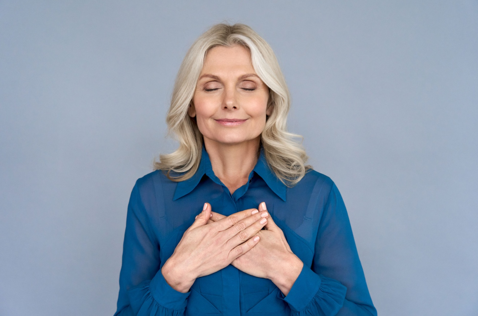 An older adult in a blue shirt showing gratitude with both eyes closed and both hands on their chest.