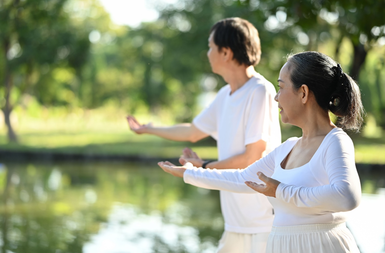 Two older adults wearing white perform balance exercises at a park.
