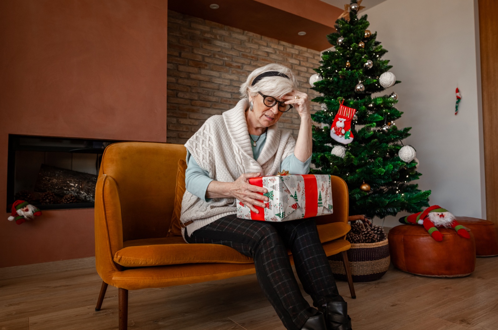 An older adult with dementia sitting on a couch with a gift on their lap with a Christmas tree behind them.