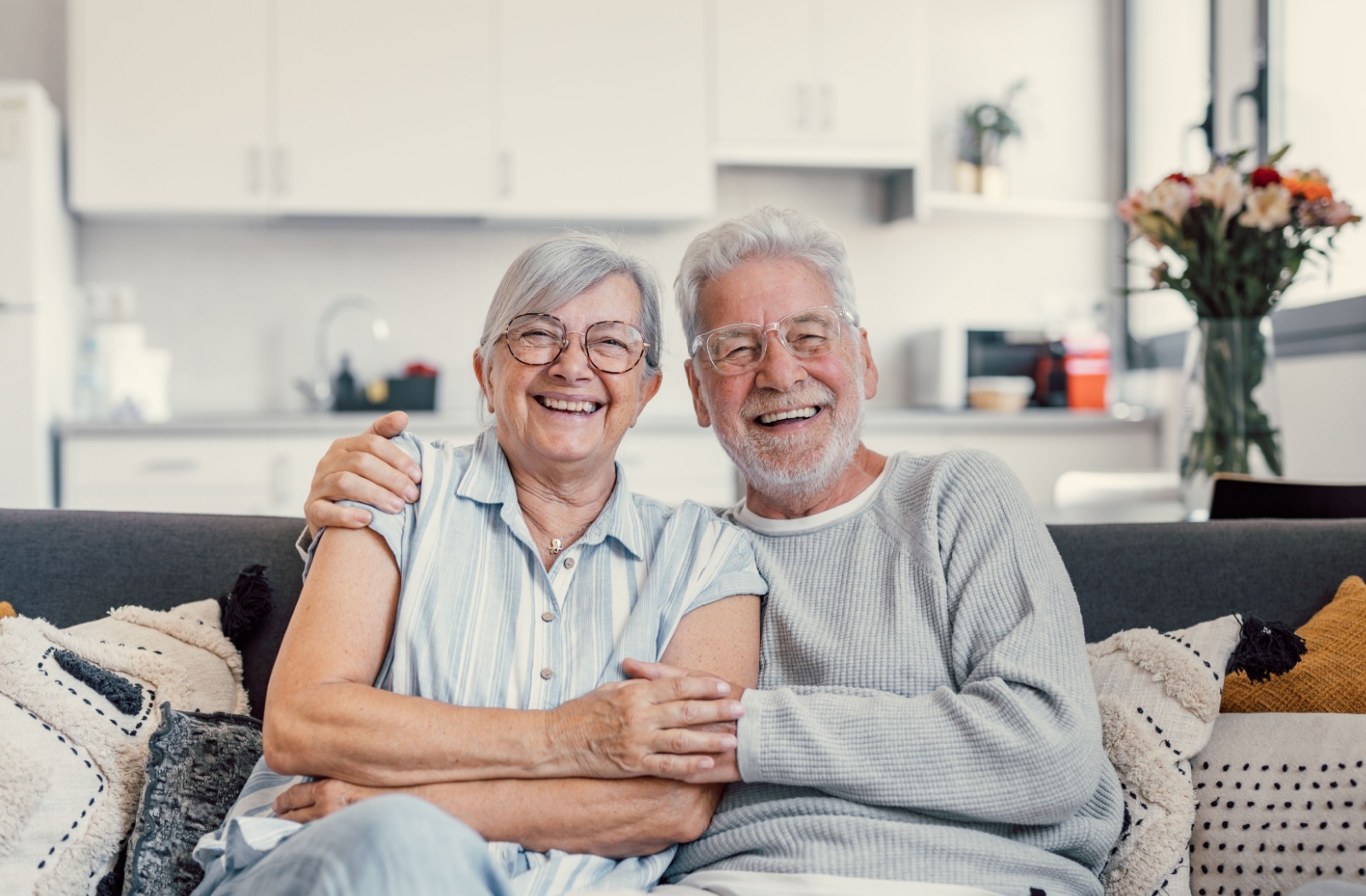 An older married couple sitting on the couch with arms around one another, smiling in their new home in independent living.