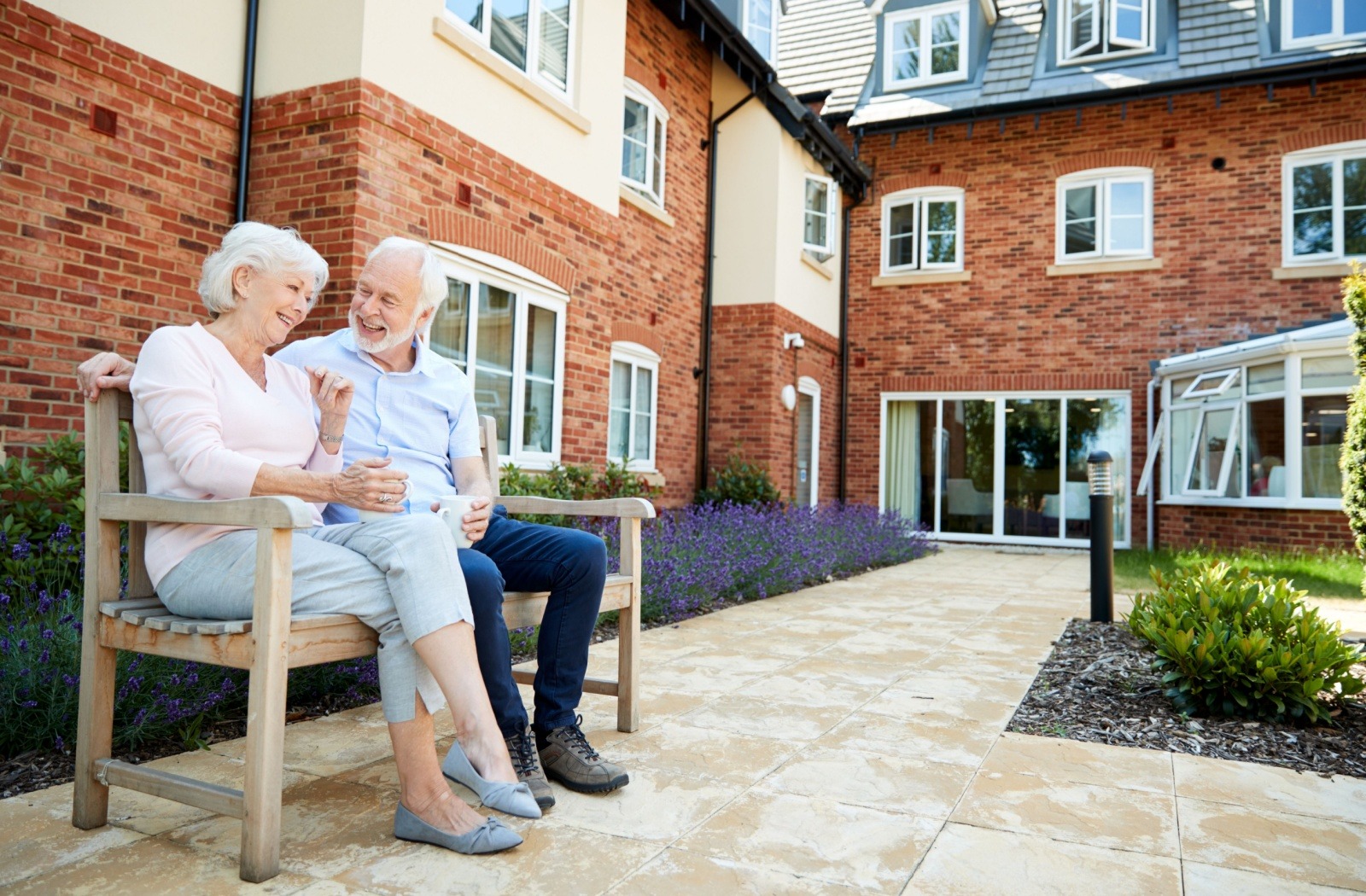 A senior couple drink coffee together while sitting on a bench outside a senior living community